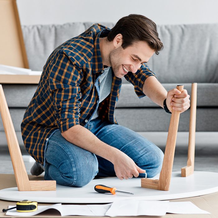 New interior furniture, home renovation process, moving, hobby and assembly table self hands. Happy millennial cute guy screwing detail with screwdriver to desk in living room interior, empty space
