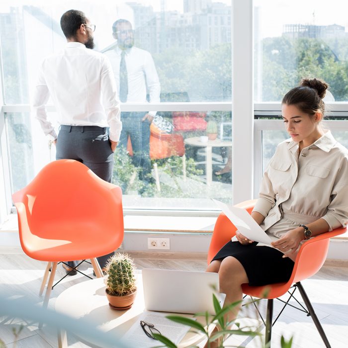 Portrait of two people in waiting room of modern office, focus on woman reading magazine in bright orange chair